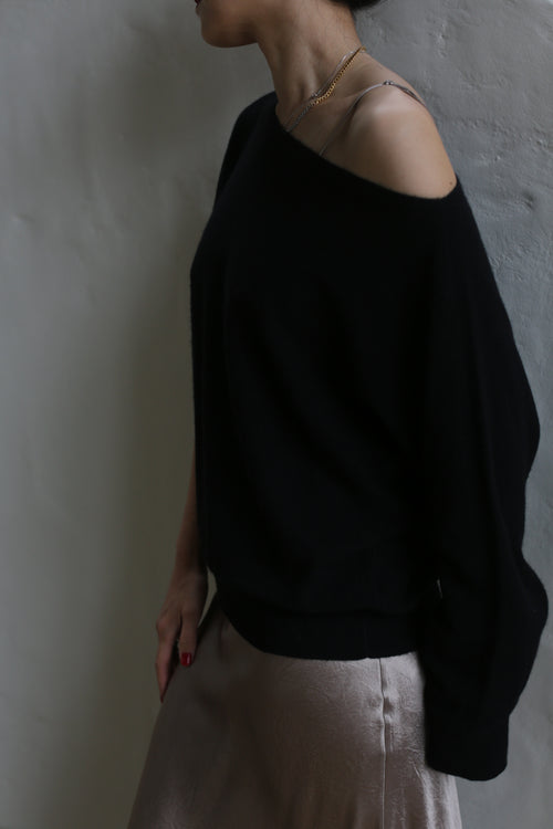 A person is standing against a plain background, wearing the Ballerina's Cashmere Sweater in black, styled off-the-shoulder, paired with a light-colored satin skirt. They are facing slightly away from the camera, with their face partially visible. The individual has one hand on their hip, exuding timeless elegance in this wardrobe staple.