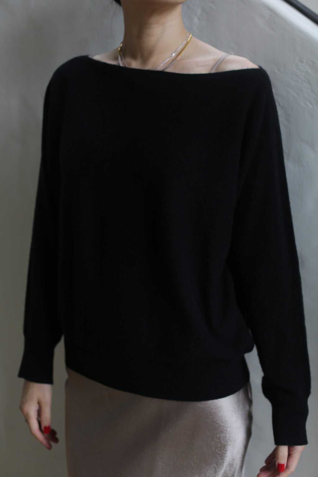 A person is wearing the Ballerina's Cashmere Sweater in black, paired with light-colored pants. Their head and lower body are out of the frame. The background is plain and slightly textured.