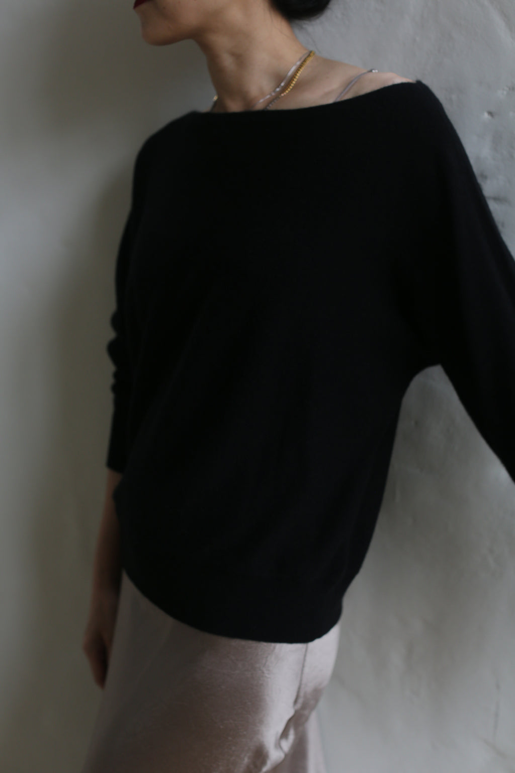 A person wearing the loose-fitting Ballerina's Cashmere Sweater in black and light-colored pants leans against a plain wall. Their face is not fully visible. The photo has a soft, slightly blurred aesthetic, capturing the timeless elegance of this wardrobe staple.