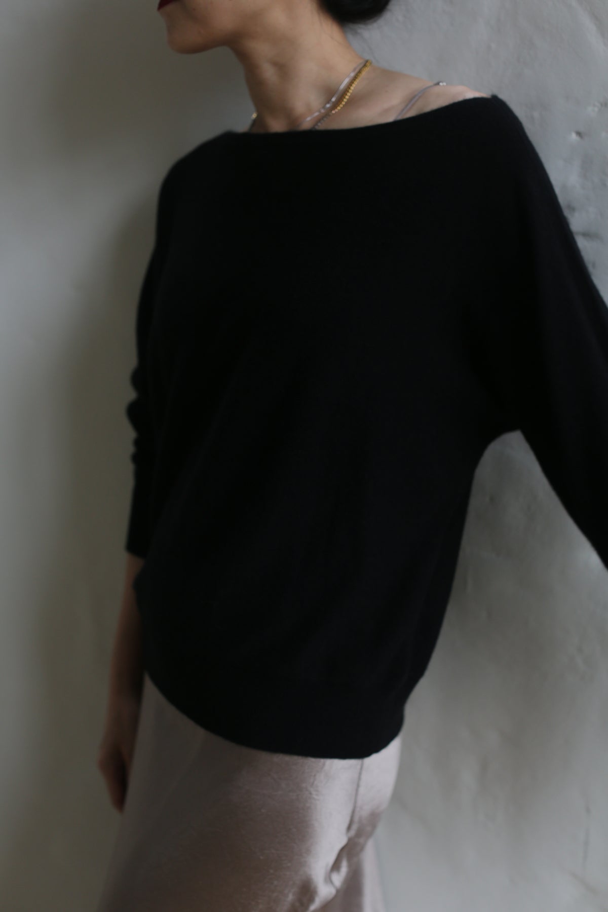 A person wearing the loose-fitting Ballerina's Cashmere Sweater in black and light-colored pants leans against a plain wall. Their face is not fully visible. The photo has a soft, slightly blurred aesthetic, capturing the timeless elegance of this wardrobe staple.