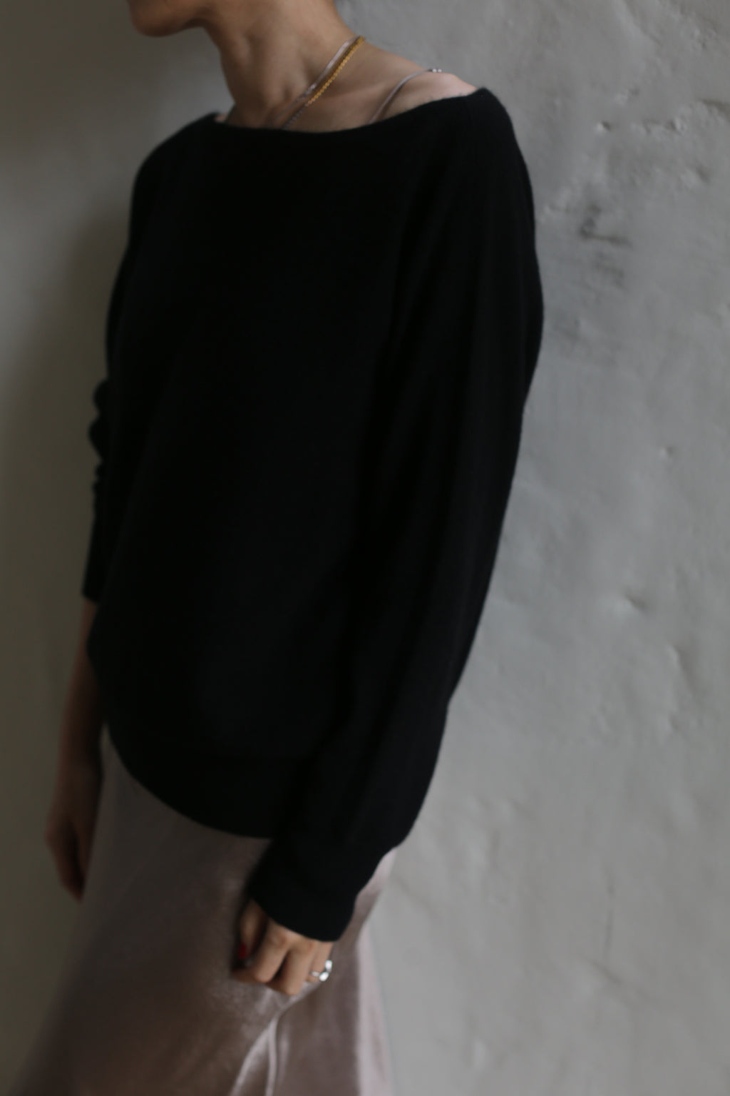 A person wearing a dark long-sleeve Ballerina's Cashmere Sweater in black and light-colored pants stands against a textured white wall. The image is slightly blurry, and the person's head is partially out of the frame.