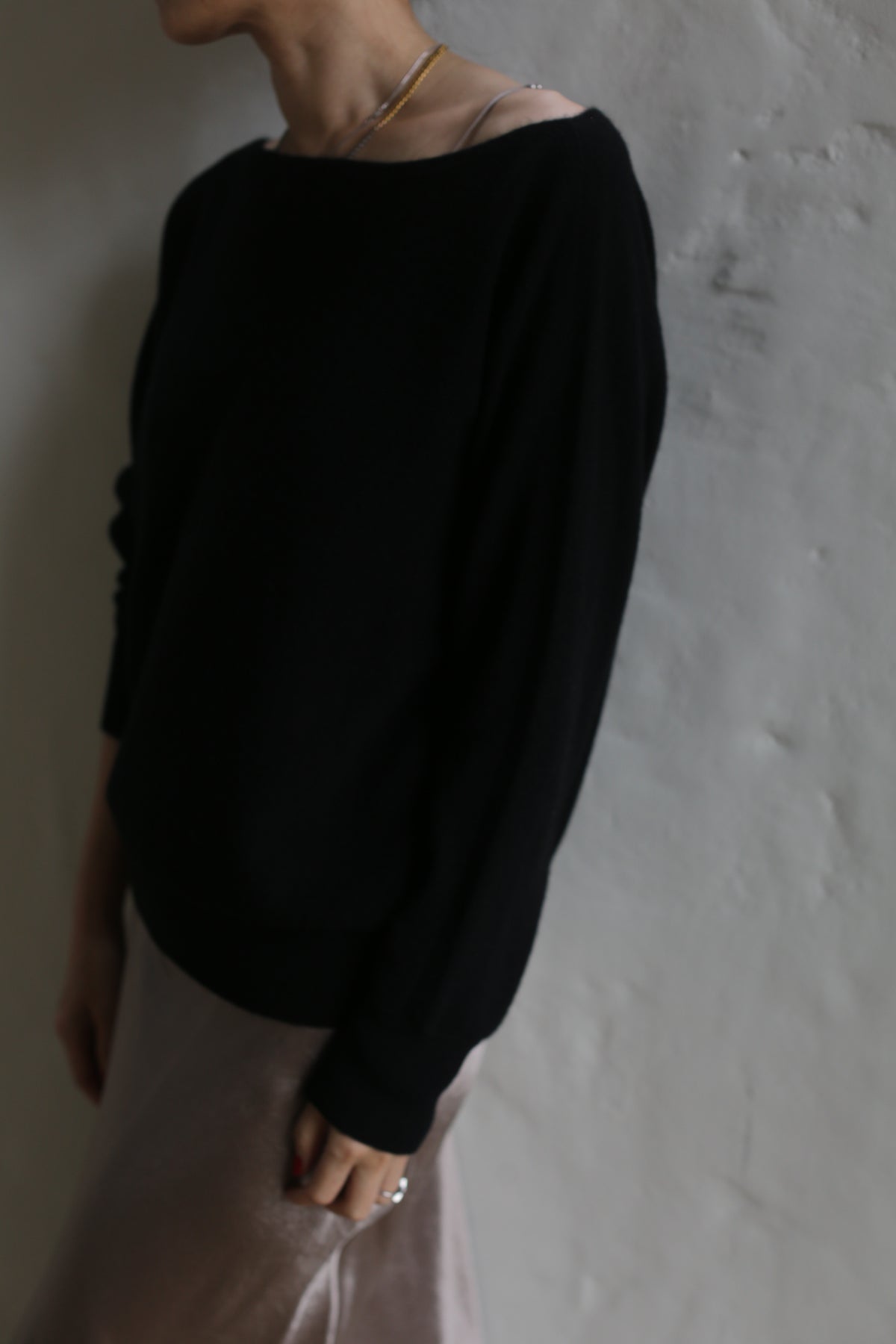A person wearing a dark long-sleeve Ballerina's Cashmere Sweater in black and light-colored pants stands against a textured white wall. The image is slightly blurry, and the person's head is partially out of the frame.