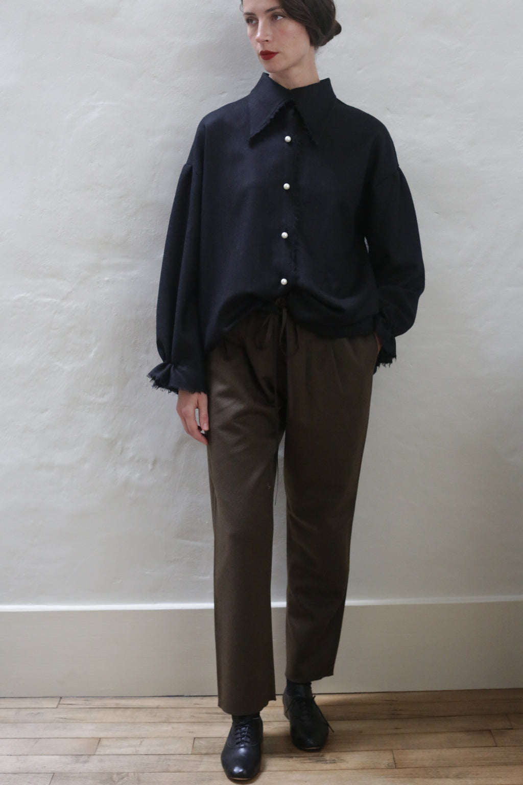 A person stands against a plain light-colored wall, wearing a loose black button-up shirt with frayed cuffs and collar, Cashmere Unisex Pants in Olive with a silk ribbon drawstring, and black shoes. They have short dark hair styled neatly, red lipstick, and their left hand in their pocket.