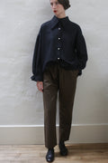 A person stands against a plain light-colored wall, wearing a loose black button-up shirt with frayed cuffs and collar, Cashmere Unisex Pants in Olive with a silk ribbon drawstring, and black shoes. They have short dark hair styled neatly, red lipstick, and their left hand in their pocket.