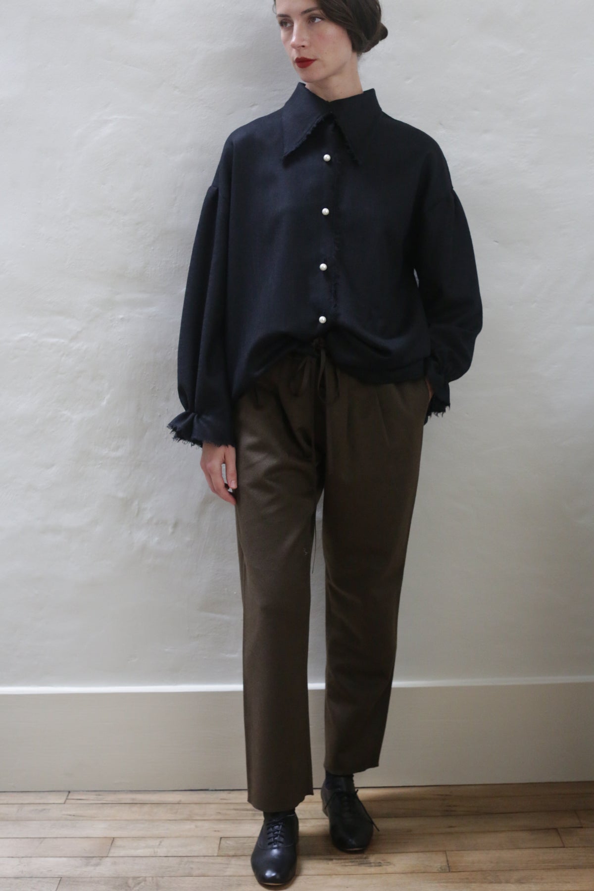 A person stands against a plain light-colored wall, wearing a loose black button-up shirt with frayed cuffs and collar, Cashmere Unisex Pants in Olive with a silk ribbon drawstring, and black shoes. They have short dark hair styled neatly, red lipstick, and their left hand in their pocket.