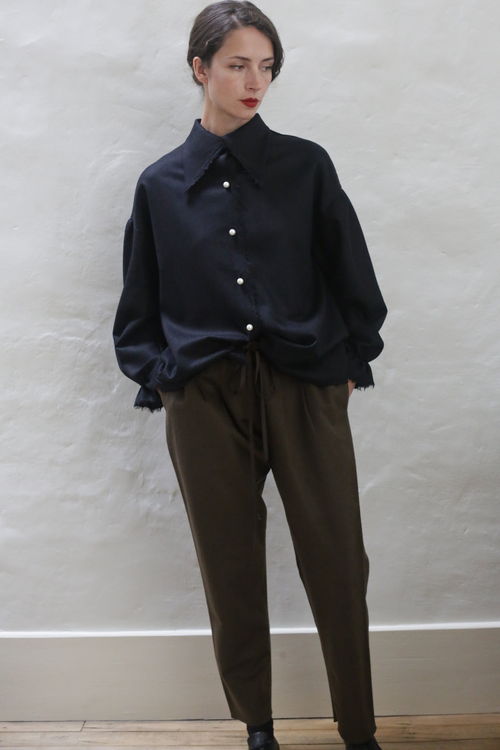 A person with short, dark hair stands against a light gray textured wall. They are wearing a loose, dark blue button-up shirt with white buttons and relaxed-fit Cashmere Unisex Pants | Olive with a silk ribbon drawstring. Their hands are in their pockets and they are wearing dark shoes. The overall look is effortlessly unisex.