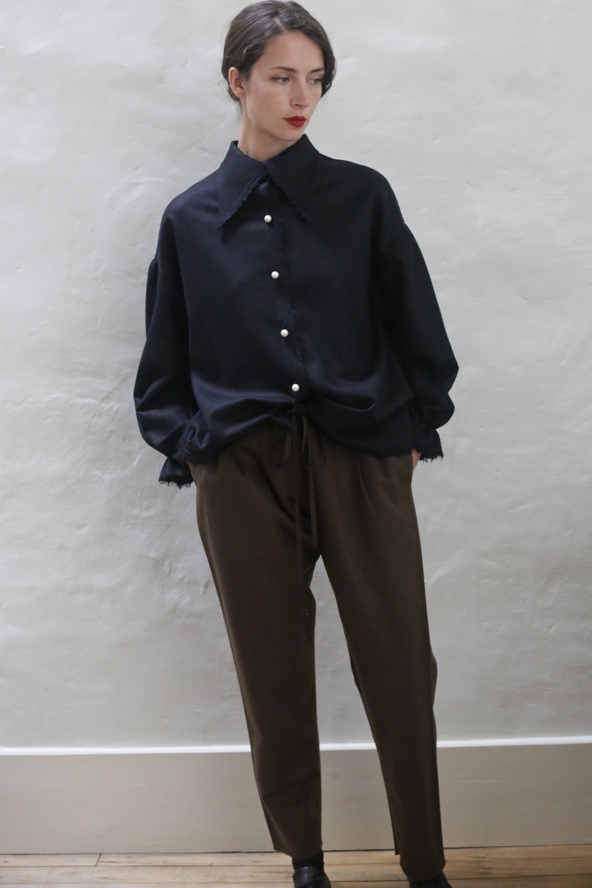 A person with short, dark hair stands against a light gray textured wall. They are wearing a loose, dark blue button-up shirt with white buttons and relaxed-fit Cashmere Unisex Pants | Olive with a silk ribbon drawstring. Their hands are in their pockets and they are wearing dark shoes. The overall look is effortlessly unisex.