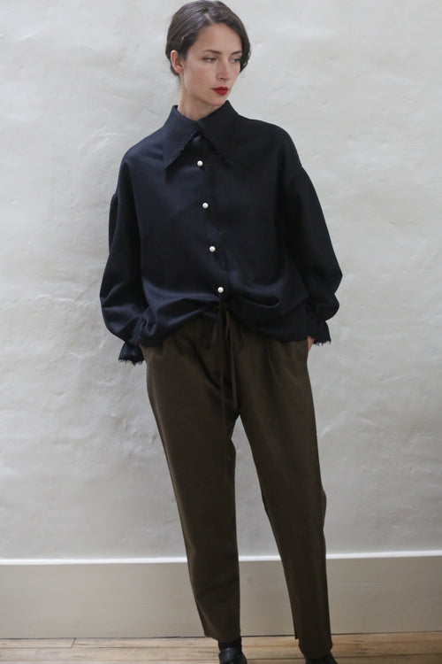 A person with short, dark hair stands against a light gray textured wall. They are wearing a loose, dark blue button-up shirt with white buttons and relaxed-fit Cashmere Unisex Pants | Olive with a silk ribbon drawstring. Their hands are in their pockets and they are wearing dark shoes. The overall look is effortlessly unisex.