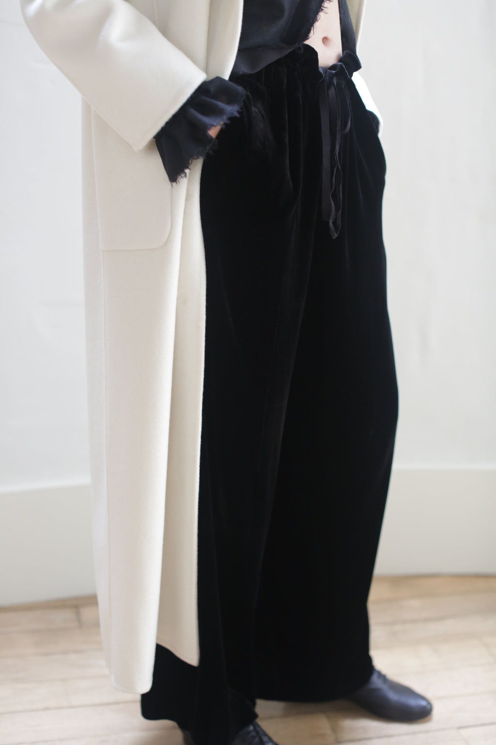 A person stands on a wooden floor, wearing a white coat over a black top and Silk Velvet Trousers tied with a ribbon. The individual's face is not visible. The outfit is completed with black shoes.