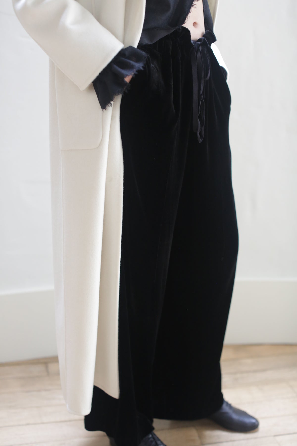 A person stands on a wooden floor, wearing a white coat over a black top and Silk Velvet Trousers tied with a ribbon. The individual's face is not visible. The outfit is completed with black shoes.