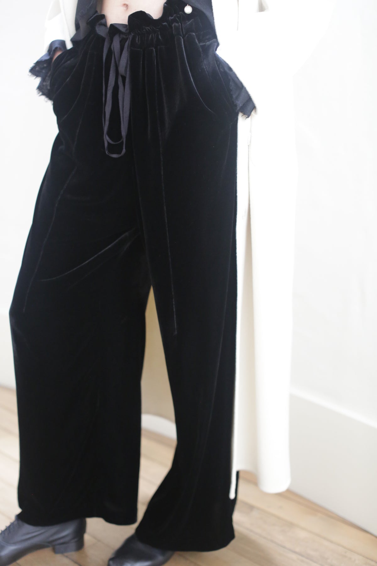 A person wears Silk Velvet Trousers paired with a black top and a white coat draped over their shoulders. The trousers are tied at the waist with a dark ribbon. They are standing on a light wooden floor.
