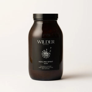 A dark brown jar with a black label prominently displaying the "Wilder Botanics" brand name. The label reads "Healing Spirit Soak," highlighting that it is calendula-infused with rose geranium, vetiver, and enriched with pink Himalayan salts. This 100% natural product stands against a light background.