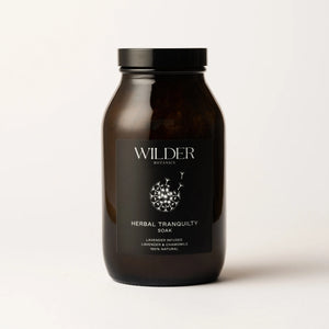 A dark brown glass jar with a black lid is labeled "Herbal Tranquility Soak" from Wilder Botanics. The label highlights that the soak is infused with lavender and soothing chamomile, complemented by Epsom salts and French green clay, ensuring a 100% natural blend. The background is plain and white.