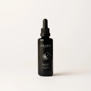 A dark-colored dropper bottle labeled "Recovery Night Oil" stands against a plain white background. The minimalist packaging features a floral design and text in white, highlighting its blend of rose oil and phytonutrients.