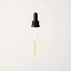 A transparent dropper featuring a black rubber top releases a single drop of vibrant yellow Phyto Radiance Day Oil against a light gray background.