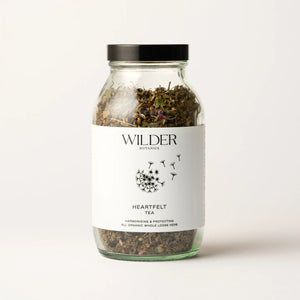 A glass jar labeled "Heartfelt Tea" contains a harmonizing herbal blend of dried herbs. The jar features a black screw-on lid and a white label adorned with a dandelion seed illustration. This organic tea, crafted for heart support with adaptogens, promises to be both harmonizing and protective.