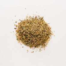 Load image into Gallery viewer, A pile of Solstice Tea is spread on a plain white surface. This herbal blend appears finely chopped with small pieces of leaves and stems visible, varying in shades of green and brown.