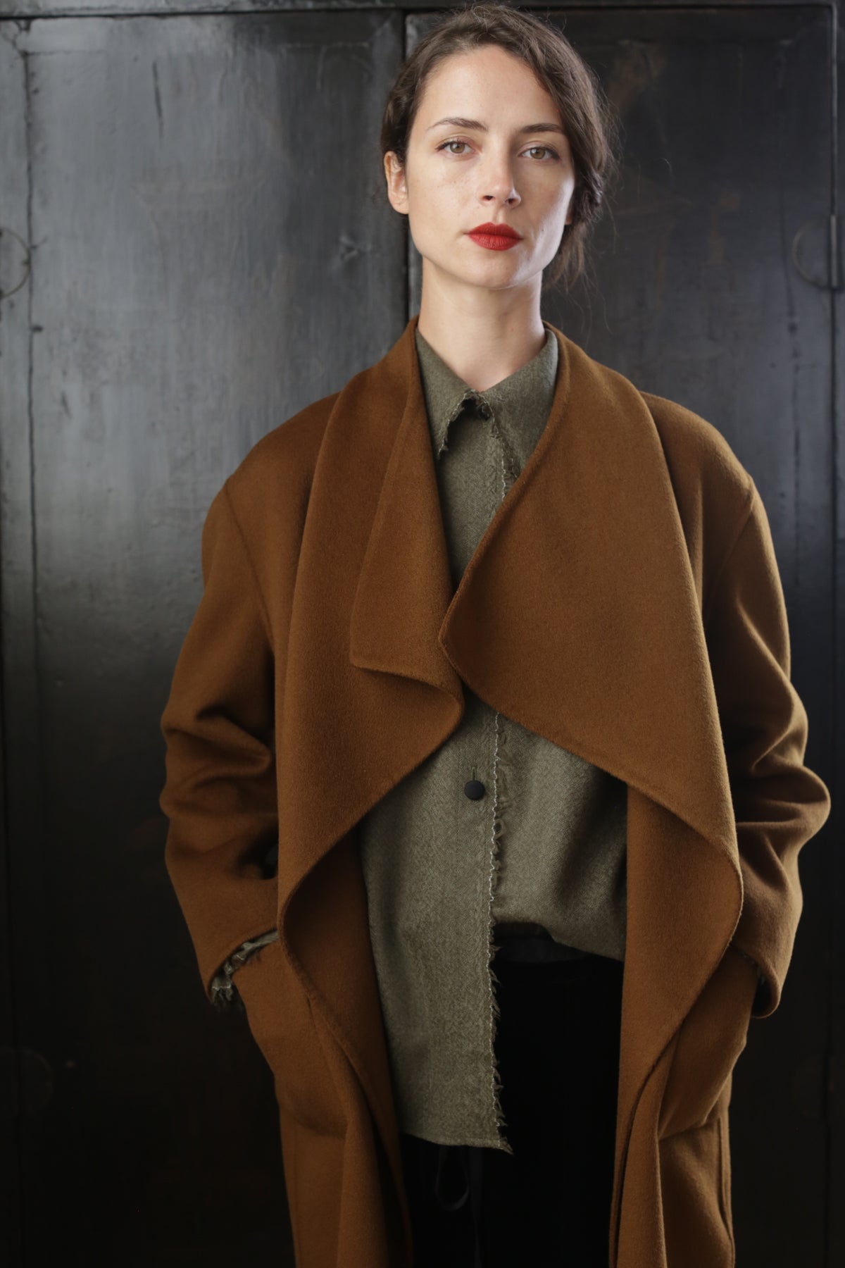 A woman stands against a dark background wearing a Winston Long Double-face and a green shirt. Her hands are in the coat pockets, and she has red lipstick on. She has a determined expression on her face, and her hair is styled in a side part.