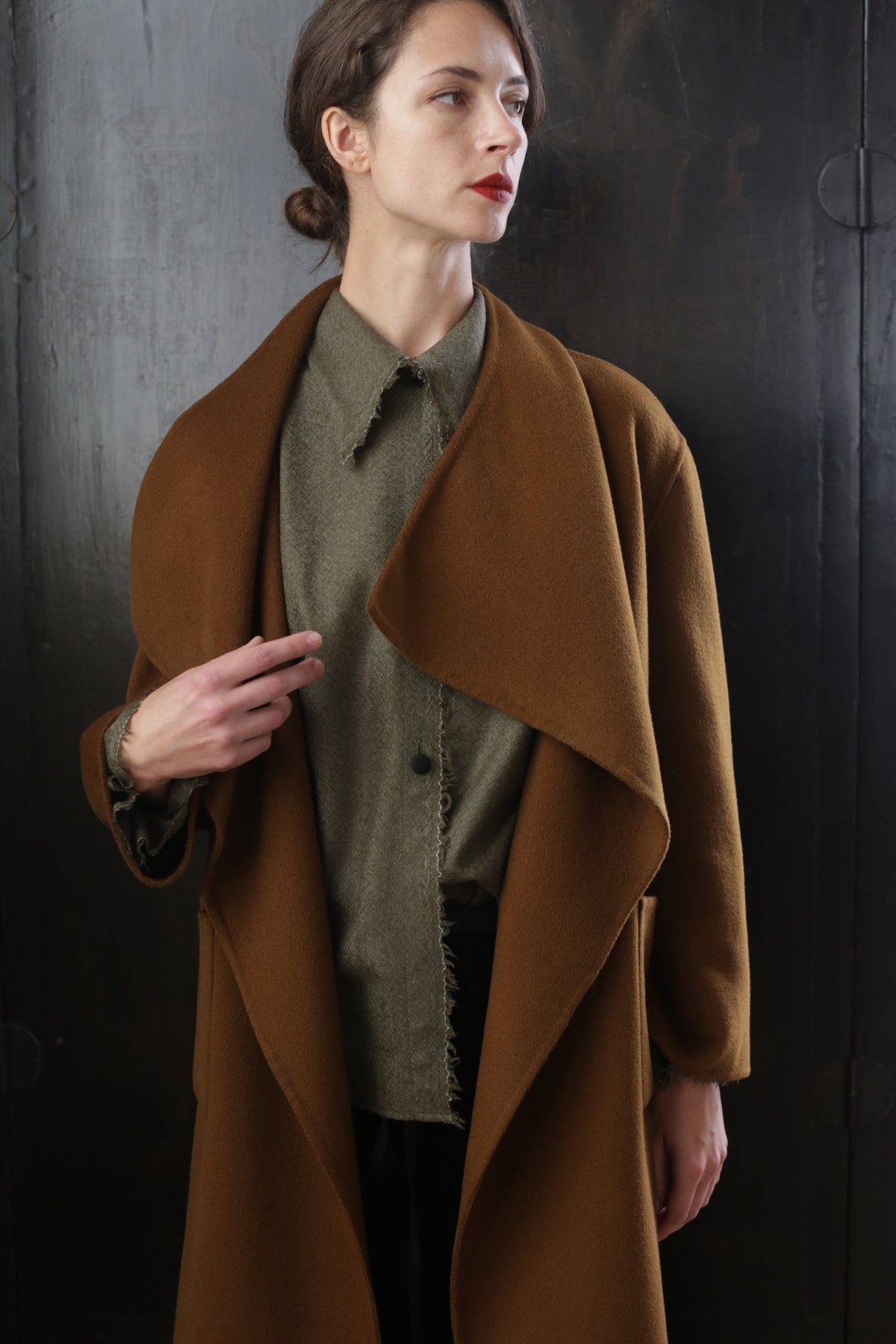 Woman with short brown hair wearing a long, tobacco-colored coat over a green collared shirt. The Winston Long Double-face showcases relaxed construction as she stands against a dark, textured background with her left hand holding the lapel. She looks slightly to the right with a neutral expression.