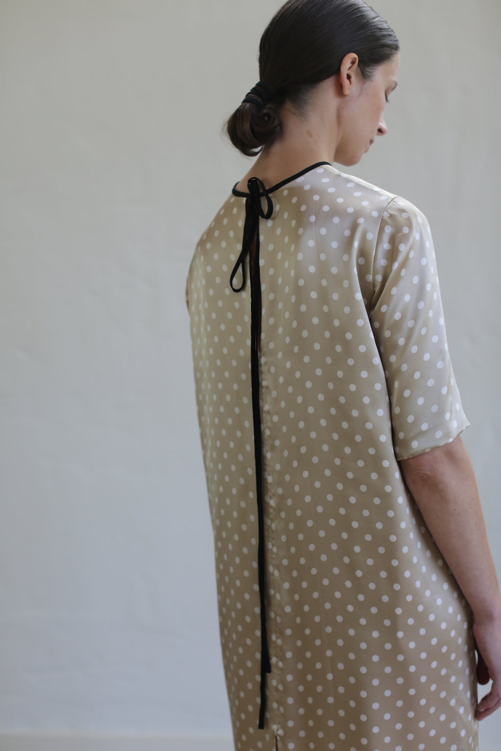 A person with their hair in a low bun is turned away, wearing a beige silk charmeuse Reversible Crewneck Dress with white polka dots. The elegant crewneck dress features a black ribbon tied at the back of the neck. The background is plain and light-colored, highlighting the sophisticated simplicity of this handmade piece from San Francisco.