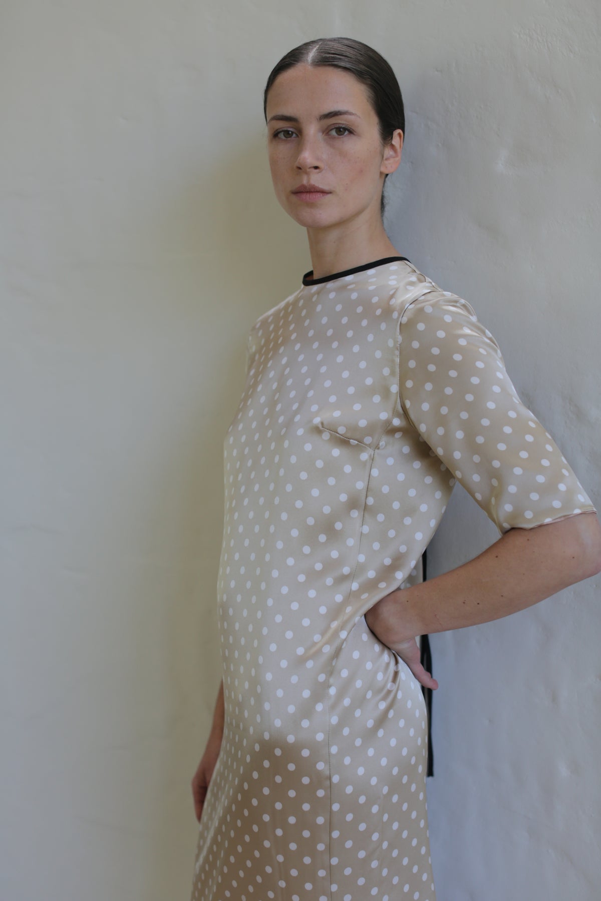 A woman with dark hair tied back stands against a light wall, wearing a beige Reversible Crewneck Dress with white polka dots. She looks directly at the camera with a composed expression, one hand resting on her hip.