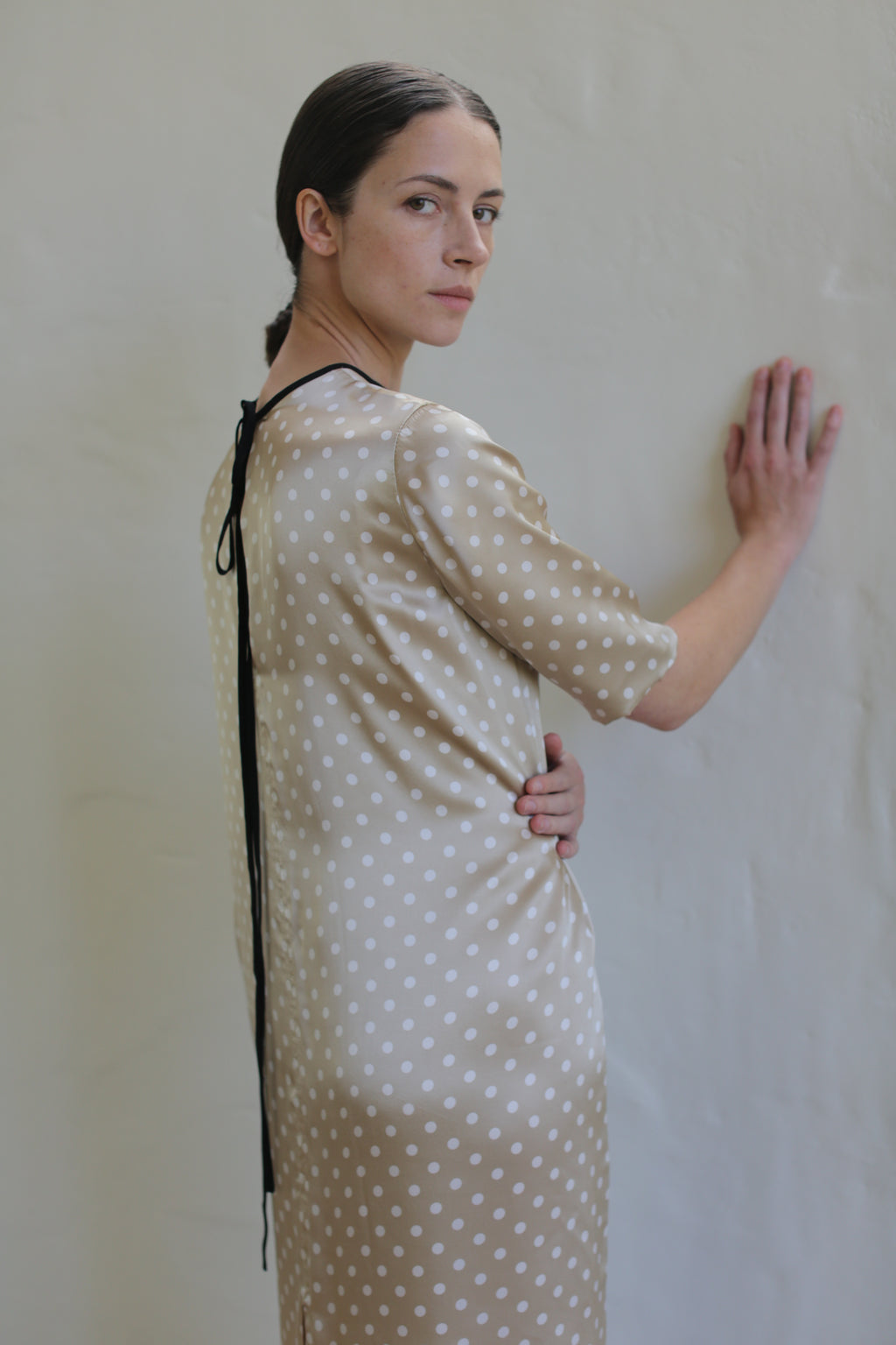 A person with long dark hair tied back stands against a light-colored wall, wearing a Reversible Crewneck Dress. They have one hand on the wall and the other on their hip, looking over their shoulder at the camera with a neutral expression.