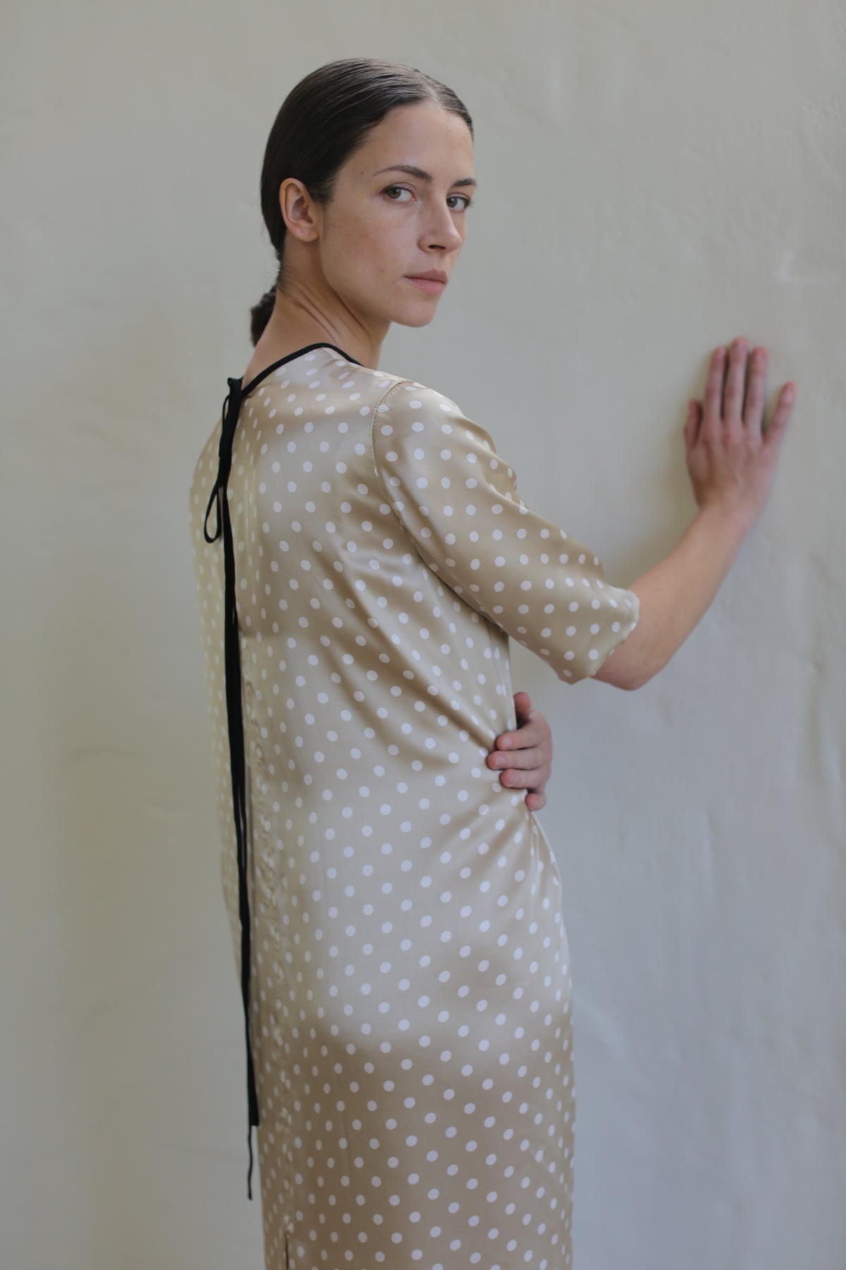 A person with long dark hair tied back stands against a light-colored wall, wearing a Reversible Crewneck Dress. They have one hand on the wall and the other on their hip, looking over their shoulder at the camera with a neutral expression.