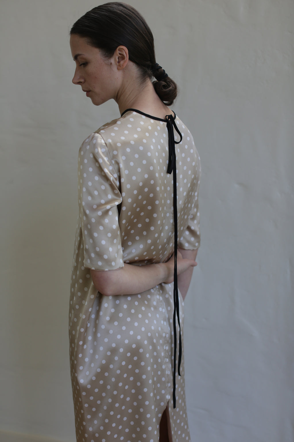 A woman with dark hair tied in a low bun is standing against a plain light background, turned sideways with her back slightly facing the camera. She is wearing a Reversible Crewneck Dress with a black ribbon tied at the neck.