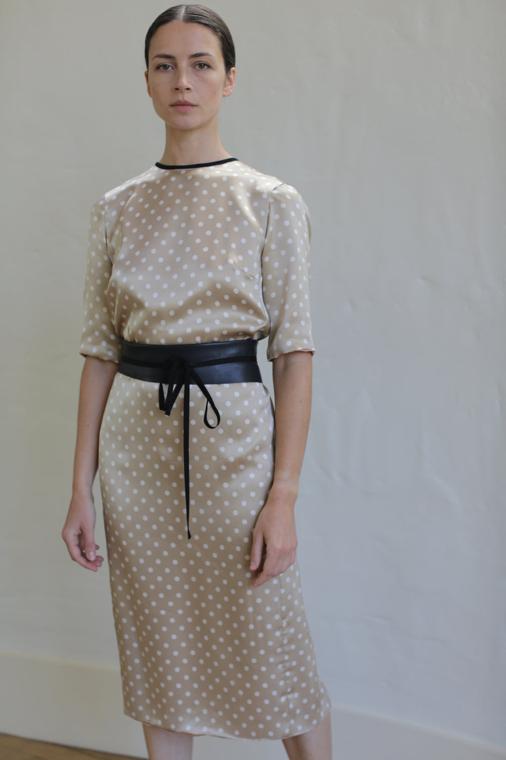 A woman stands against a plain background, wearing a beige Reversible Crewneck Dress with white polka dots. The dress, handmade in San Francisco from silk charmeuse, features elbow-length sleeves and a black belt tied at the waist. She has dark hair pulled back and maintains a neutral expression on her face.