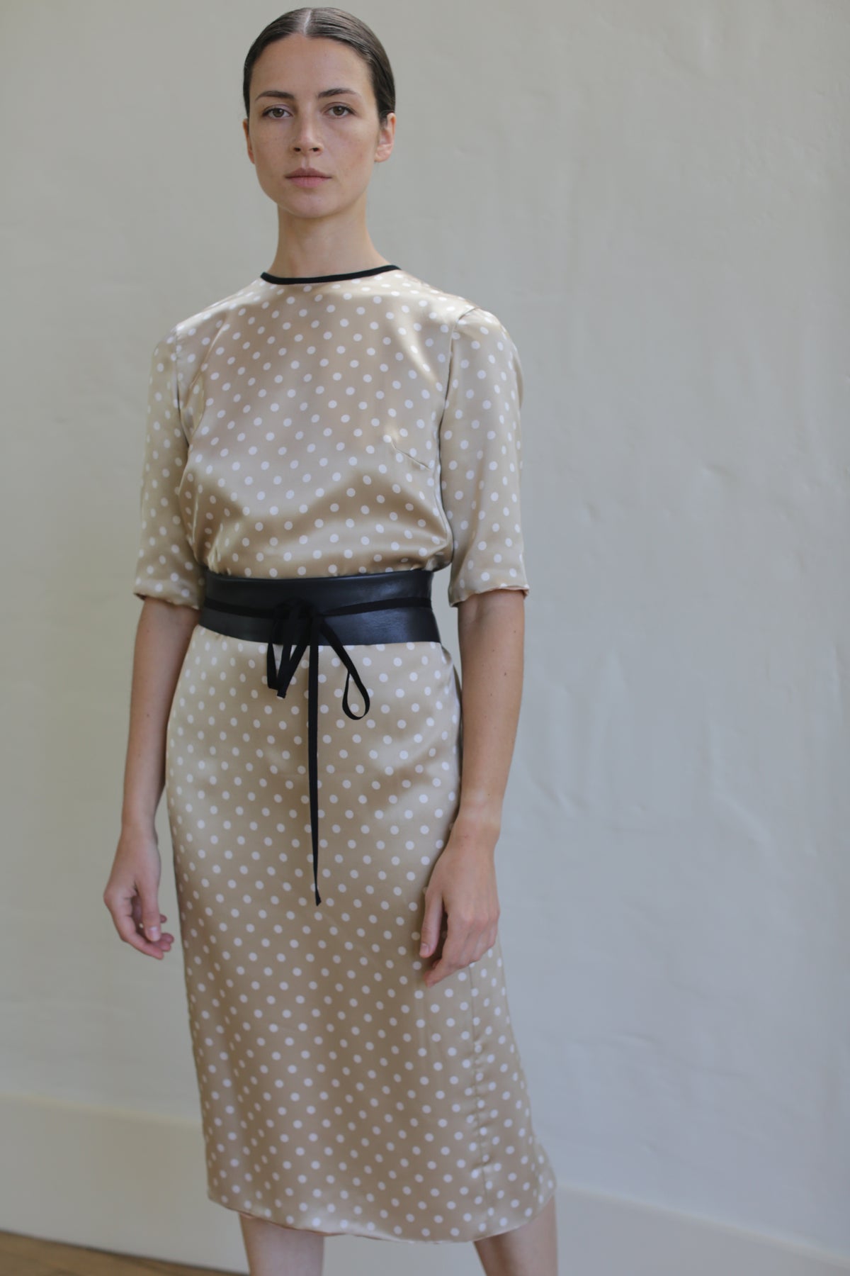 A woman stands against a plain background, wearing a beige Reversible Crewneck Dress with white polka dots. The dress, handmade in San Francisco from silk charmeuse, features elbow-length sleeves and a black belt tied at the waist. She has dark hair pulled back and maintains a neutral expression on her face.