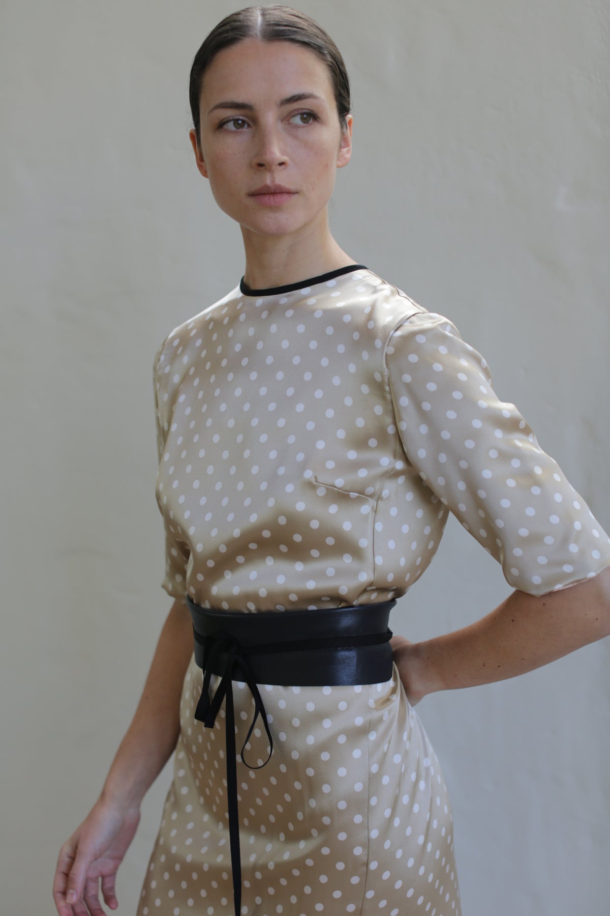 A person with dark hair tied back wears a beige Reversible Crewneck Dress with white polka dots and a black belt. Handmade in San Francisco, the outfit stands out against a plain light-colored background as they look slightly to the side.