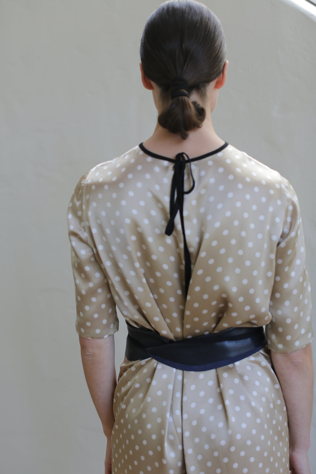 A person with dark, neatly tied hair faces away from the camera, wearing a beige dress with white polka dots. The Reversible Crewneck Dress features a black ribbon tie at the neckline and a black belt at the waist. Handmade in San Francisco, it stands out against the plain, light-colored wall background.