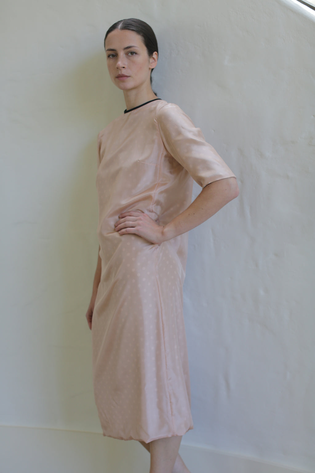 A person stands against a light-colored wall wearing a handmade in San Francisco pink, mid-length, short-sleeved Reversible Crewneck Dress. They have one hand on their hip and look directly at the camera with a neutral expression. The crewneck dress features a subtle polka dot pattern and a black neckline trim.