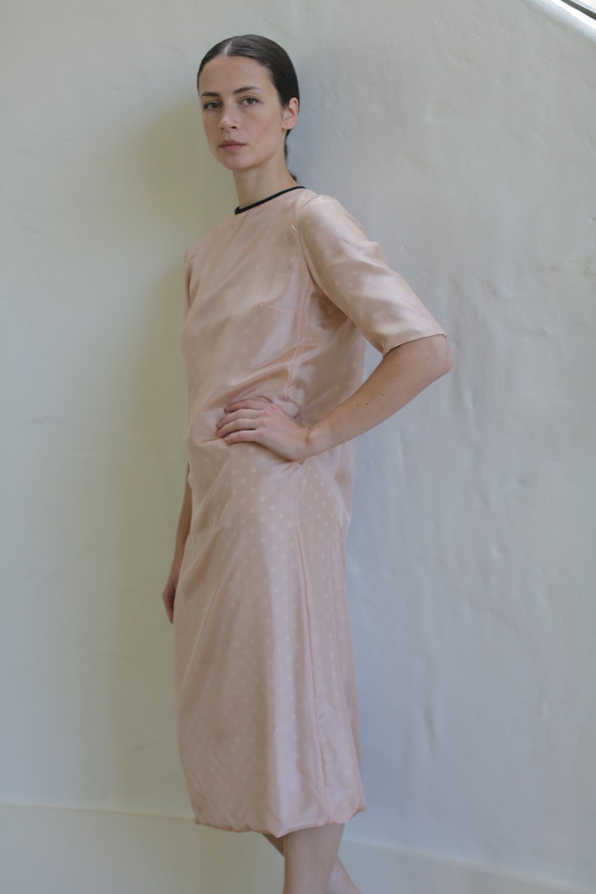 A person stands against a light-colored wall wearing a handmade in San Francisco pink, mid-length, short-sleeved Reversible Crewneck Dress. They have one hand on their hip and look directly at the camera with a neutral expression. The crewneck dress features a subtle polka dot pattern and a black neckline trim.