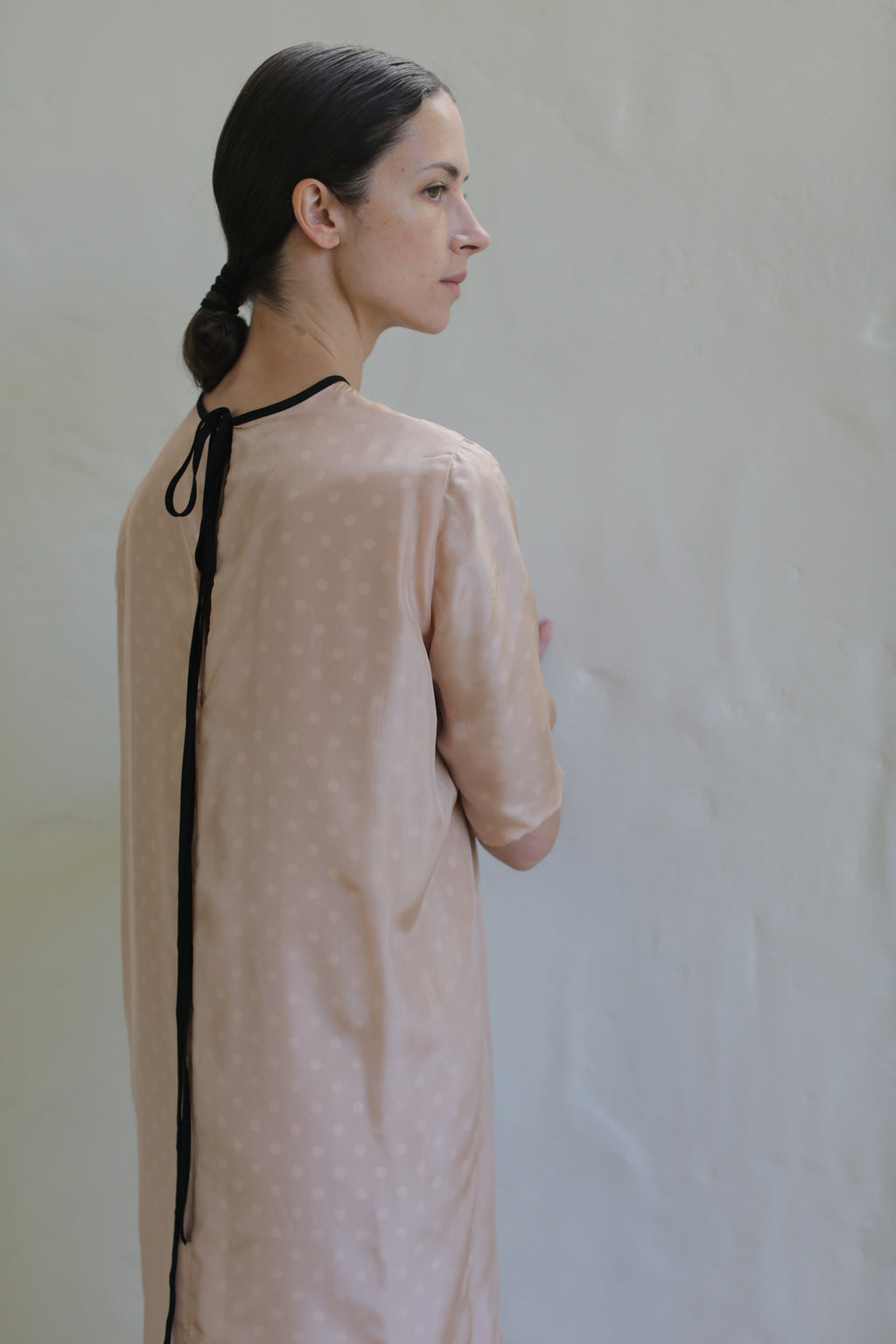 A woman wearing a light pink Reversible Crewneck Dress with subtle polka dots stands sideways, facing a plain light-colored wall. Her dark hair is neatly tied back in a low ponytail with a black hair tie that has long ends hanging down her back.