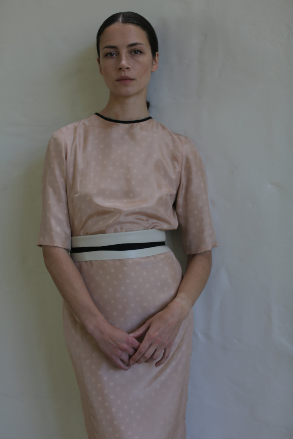 A woman stands against a light beige wall, wearing a handmade in San Francisco, knee-length, short-sleeved Reversible Crewneck Dress with subtle polka dots. She has her hair pulled back and her hands clasped in front of her. The dress features a slim white and black belt at the waist.