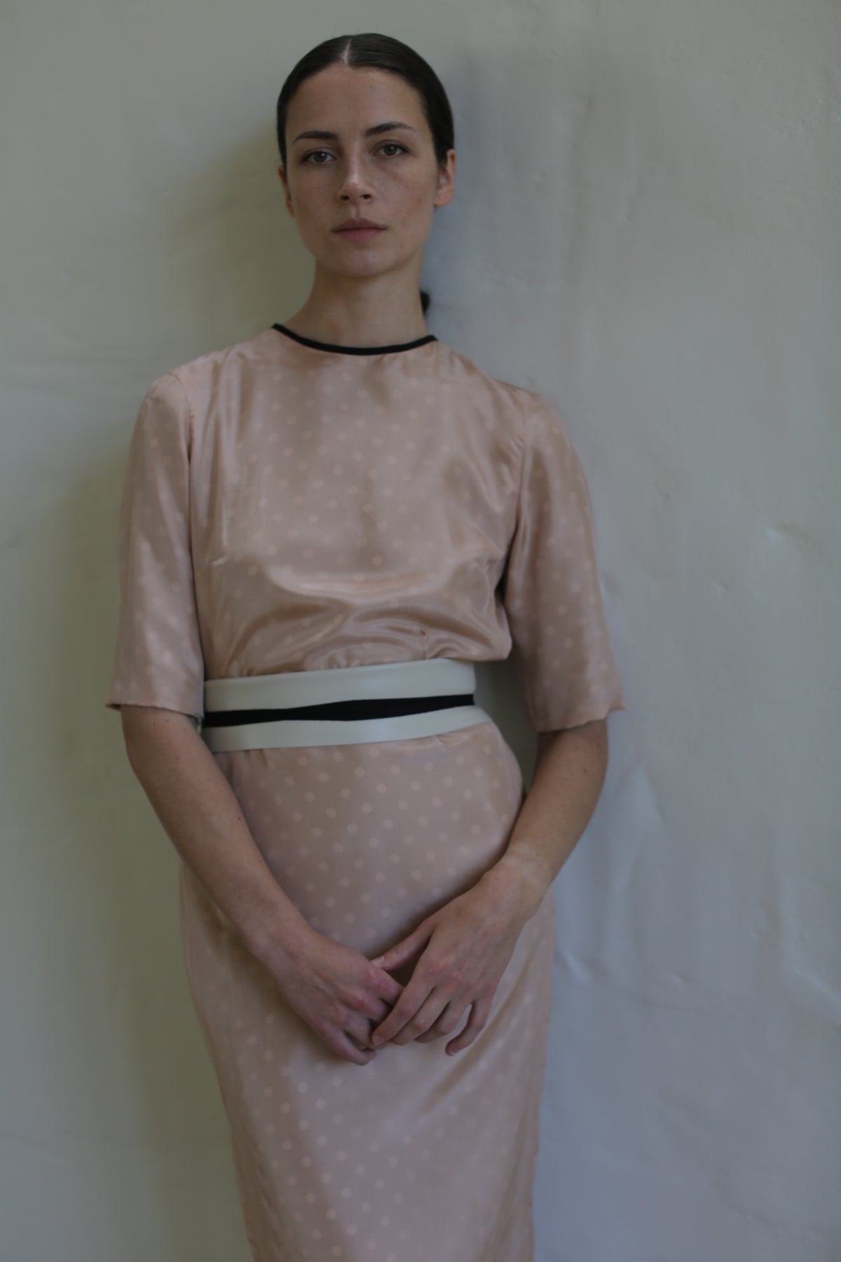 A woman stands against a light beige wall, wearing a handmade in San Francisco, knee-length, short-sleeved Reversible Crewneck Dress with subtle polka dots. She has her hair pulled back and her hands clasped in front of her. The dress features a slim white and black belt at the waist.