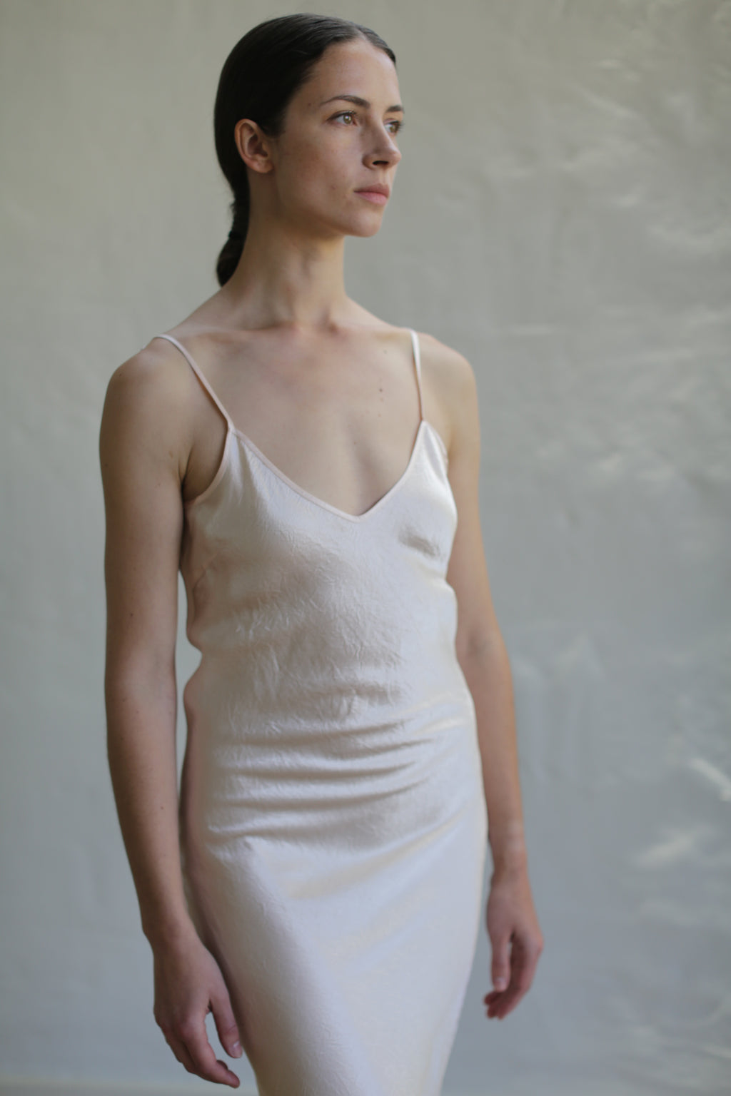 A woman with dark hair tied back gazes off to the side while wearing a Liquid Slip Dress | Champagne made from 100% plant-derived material. She stands against a plain, light background, conveying a contemplative expression.