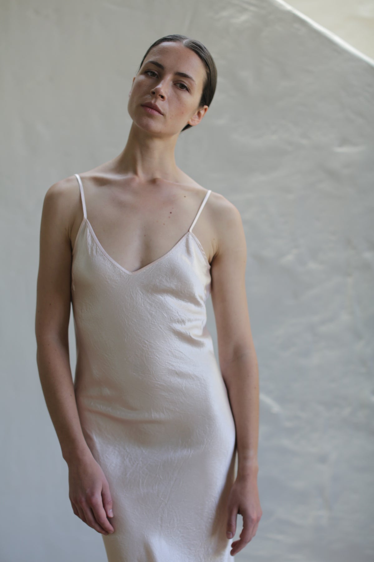 A person with long dark hair tied back stands against a plain, light-colored background, wearing a Liquid Slip Dress | Champagne made from 100% plant-derived material. The light-colored, sleeveless dress with thin straps enhances their elegant look. With a calm expression and head slightly tilted to one side, they exude serenity.