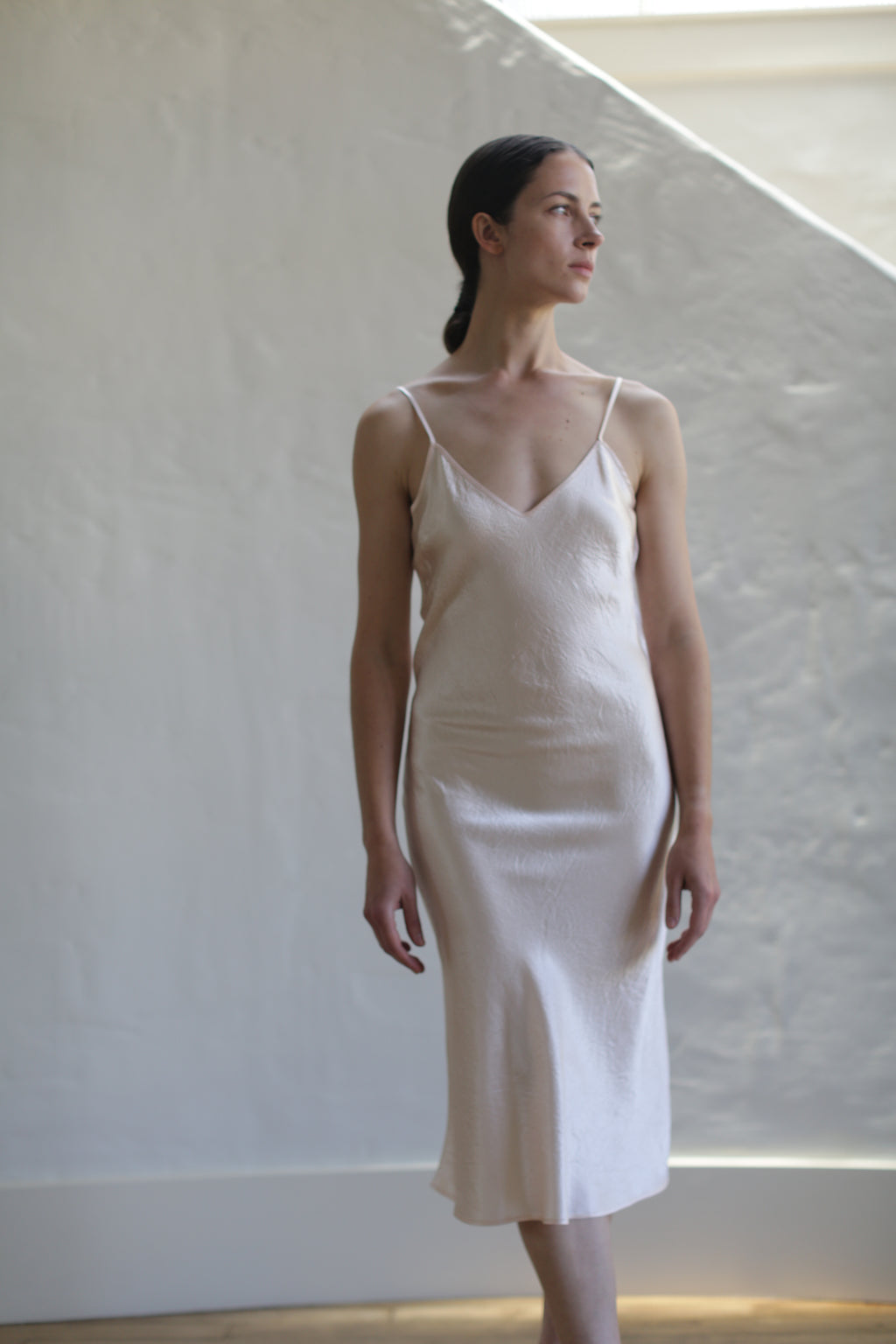 A woman stands in a minimalist setting wearing a Liquid Slip Dress | Champagne made from 100% plant-derived material. She has her hair tied back and gazes to the side. The background features a textured, neutral-toned wall with soft lighting, highlighting the elegance of her biodegradable attire.
