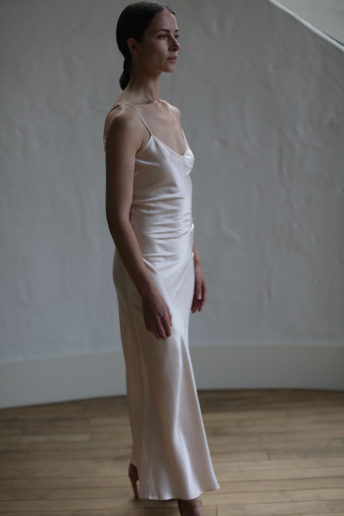 A woman with long dark hair is standing on a wooden floor wearing a Liquid Slip Dress Long | Champagne made from plant-derived material. The sexy dress has thin straps and flows down to her ankles. She is looking to the side, and the background is a textured white wall.
