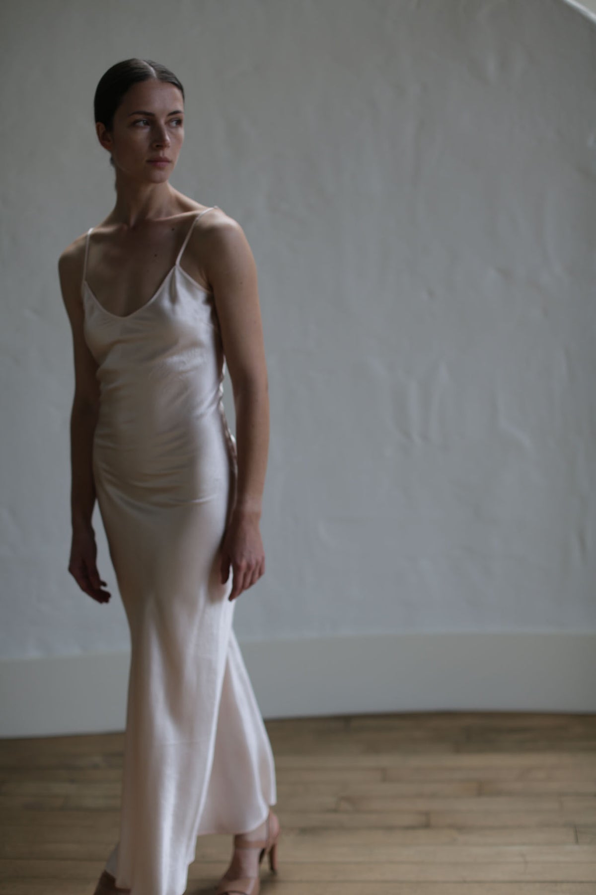 A woman stands in a softly lit room wearing a Liquid Slip Dress Long | Champagne made from plant-derived material. She looks to the side with a calm expression. The floor is wooden, and the background is a plain, textured light wall. The overall mood is serene and elegant.