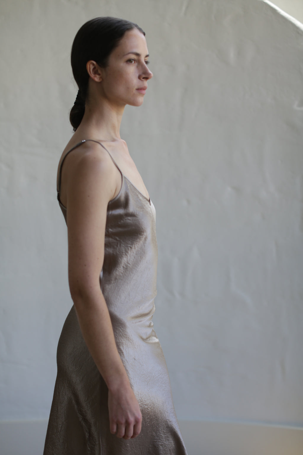 A person with dark hair, pulled back, wearing a Liquid Slip Dress | Mink made from plant-derived materials stands in profile against a neutral, textured background. The individual looks straight ahead, emanating a calm and contemplative demeanor.