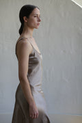 A person with dark hair, pulled back, wearing a Liquid Slip Dress | Mink made from plant-derived materials stands in profile against a neutral, textured background. The individual looks straight ahead, emanating a calm and contemplative demeanor.