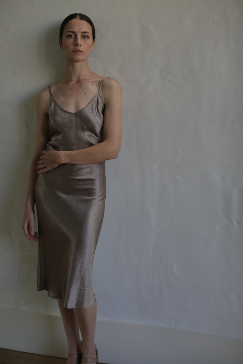 A woman with dark hair tied back is standing against a textured white wall, wearing a simple, elegant Liquid Slip Dress | Mink made of plant-derived material that reaches below her knees. Her gaze is directed toward the camera, and one hand rests on her waist while the other hangs by her side.