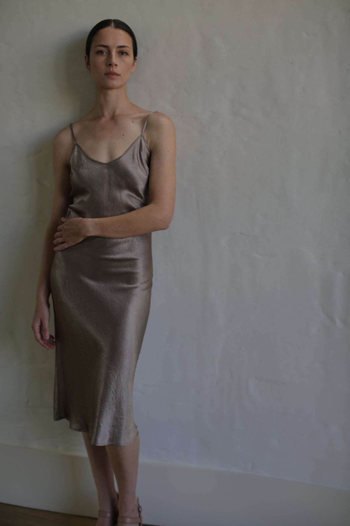 A woman with dark hair tied back is standing against a textured white wall, wearing a simple, elegant Liquid Slip Dress | Mink made of plant-derived material that reaches below her knees. Her gaze is directed toward the camera, and one hand rests on her waist while the other hangs by her side.
