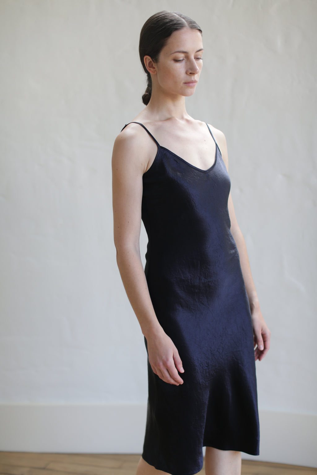 A woman with closed eyes stands against a textured white wall, wearing the Liquid Slip Dress | Midnight—a simple, sleeveless black design with thin straps. Her hair is pulled back and her hands rest by her sides. The setting is softly lit.