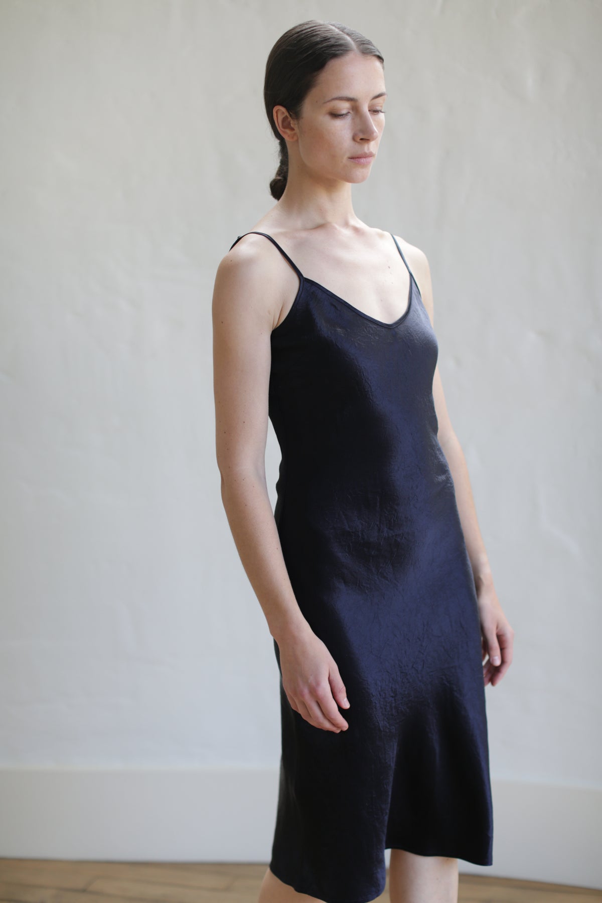 A woman with closed eyes stands against a textured white wall, wearing the Liquid Slip Dress | Midnight—a simple, sleeveless black design with thin straps. Her hair is pulled back and her hands rest by her sides. The setting is softly lit.