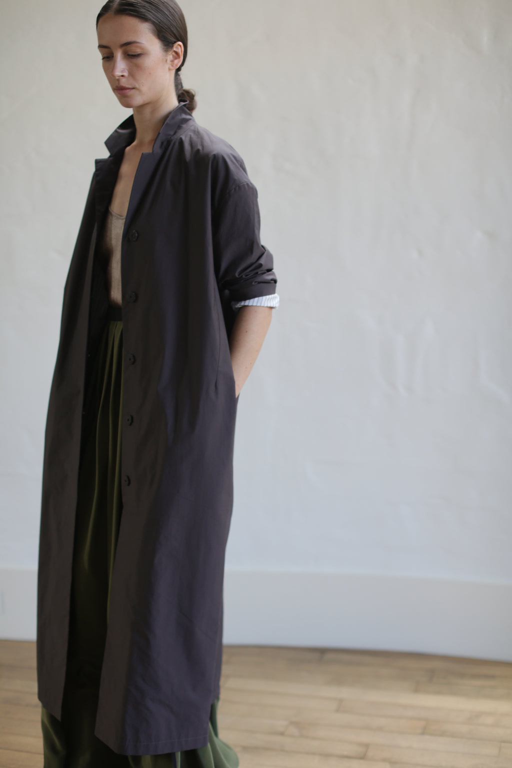 A person stands indoors wearing a brown Painter's Coat over a green skirt, accented by a leather Obi belt. They have one hand in their pocket and gaze downward. The background is a plain, light-colored wall and wooden floor.