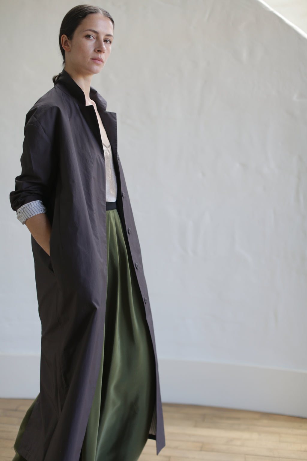 A person stands confidently in a long brown Painter's Coat with one hand in a pocket. They wear a green skirt cinched by a leather Obi Belt and pair it with a light-colored top, posing against a plain cream background.